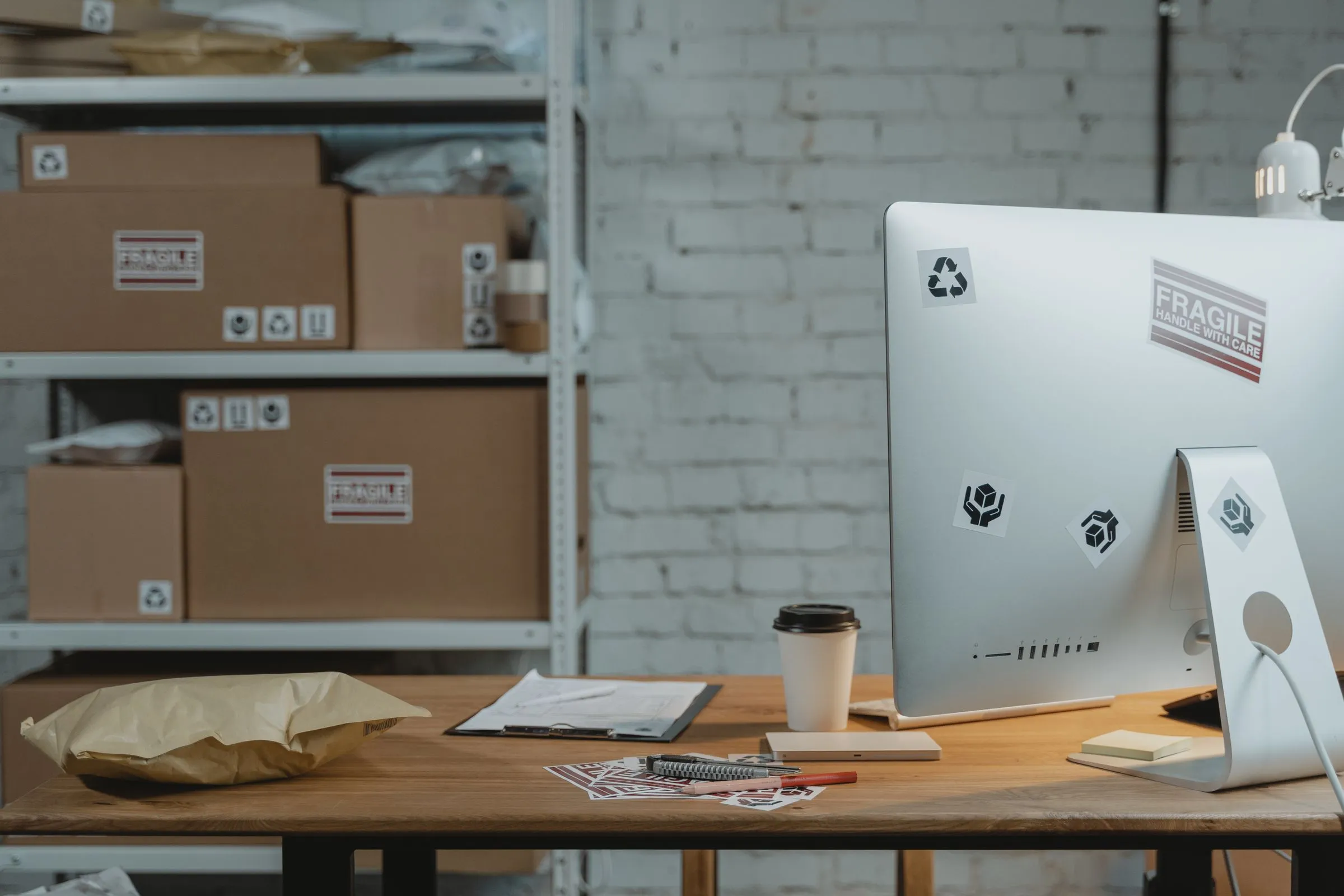 A wooden desk in a small brand&rsquo;s stockroom: a desktop computer covered in shipping and recycling stickers, a clipboard with a printed form, a kraft paper mailer, pens and a takeaway coffee cup, with a white steel shelving unit of labelled cardboard cartons against a whitewashed brick wall behind.