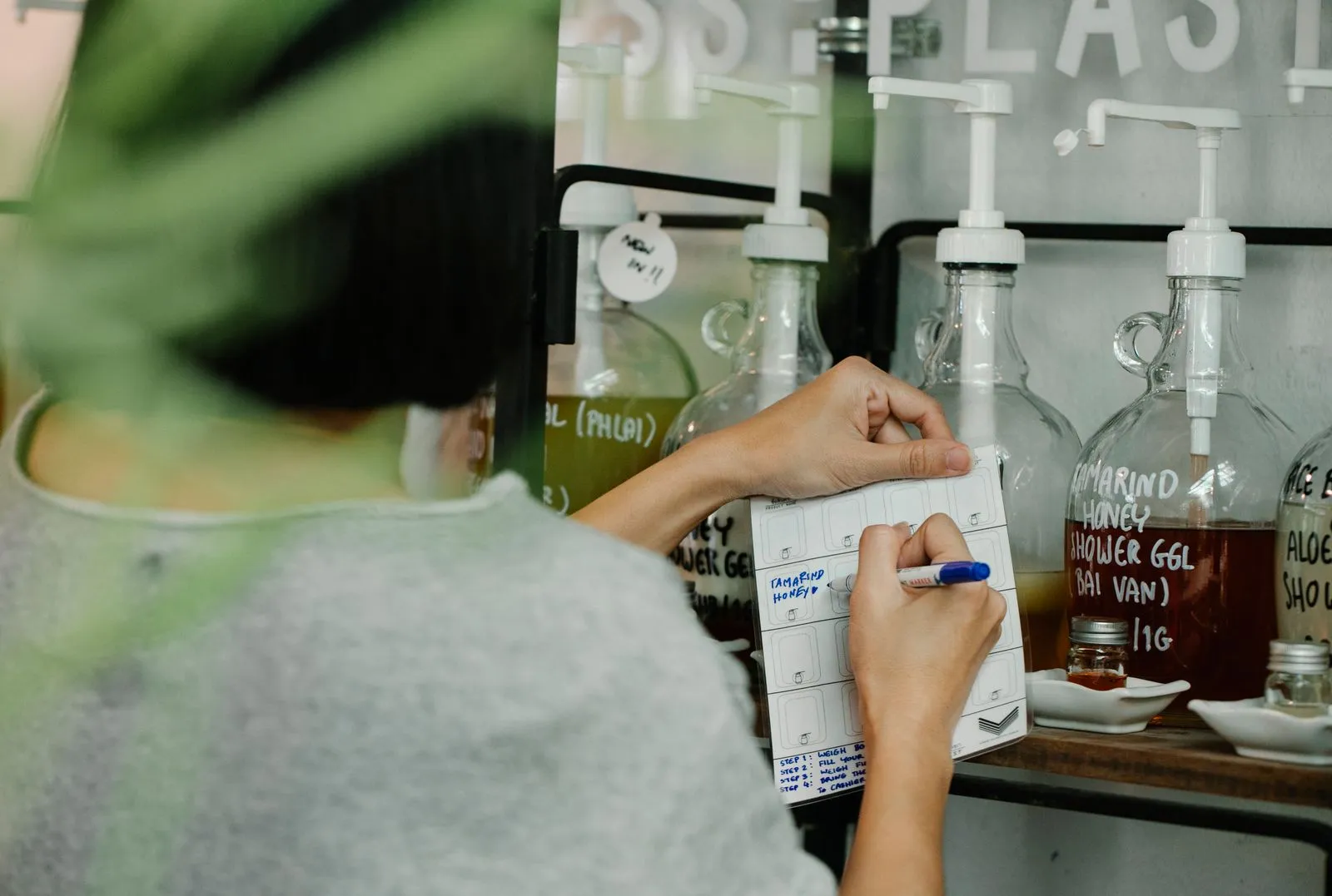 A shop worker seen from behind writes a product name onto a printed tally sheet while standing at a refill station lined with large labelled glass dispenser bottles.