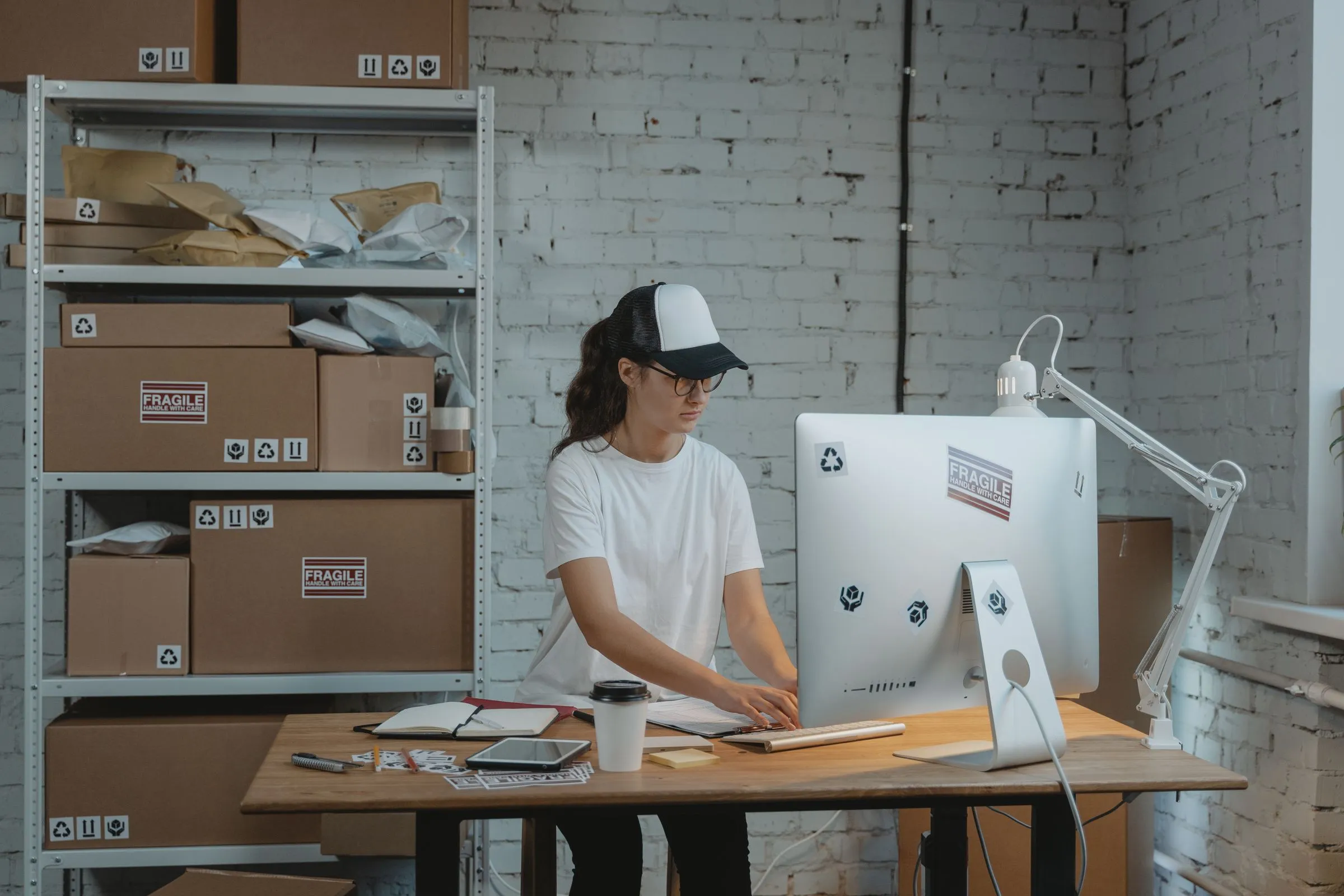 A woman in a white T-shirt and a black-and-white cap standing at a wooden desk in a stockroom, working at a desktop computer, with an open notebook, a tablet and a coffee cup on the desk and a white steel shelving unit of labelled cardboard cartons and poly mailers to her left.