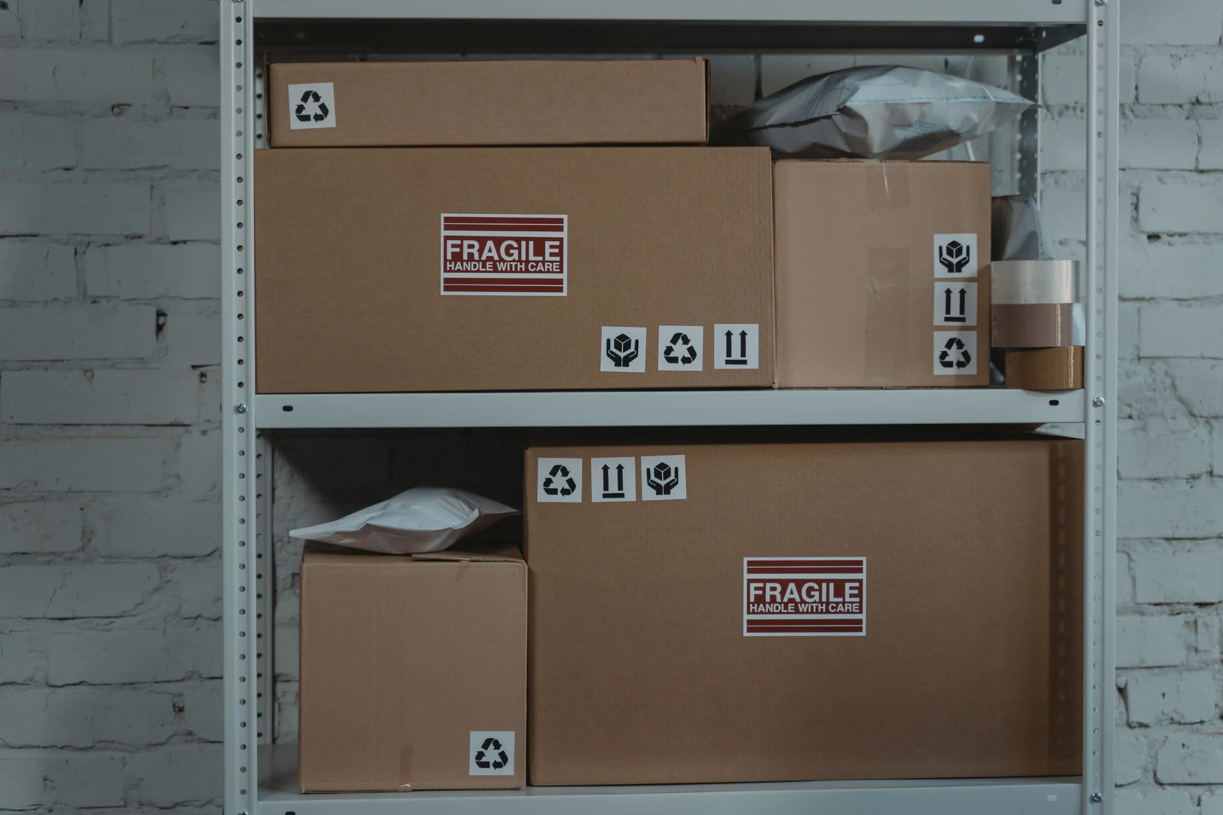 Two shelves of a white steel storage rack holding flat and square kraft cardboard cartons marked &ldquo;Fragile &mdash; handle with care&rdquo;, alongside white poly mailer bags and a roll of packing tape, against a whitewashed brick wall.